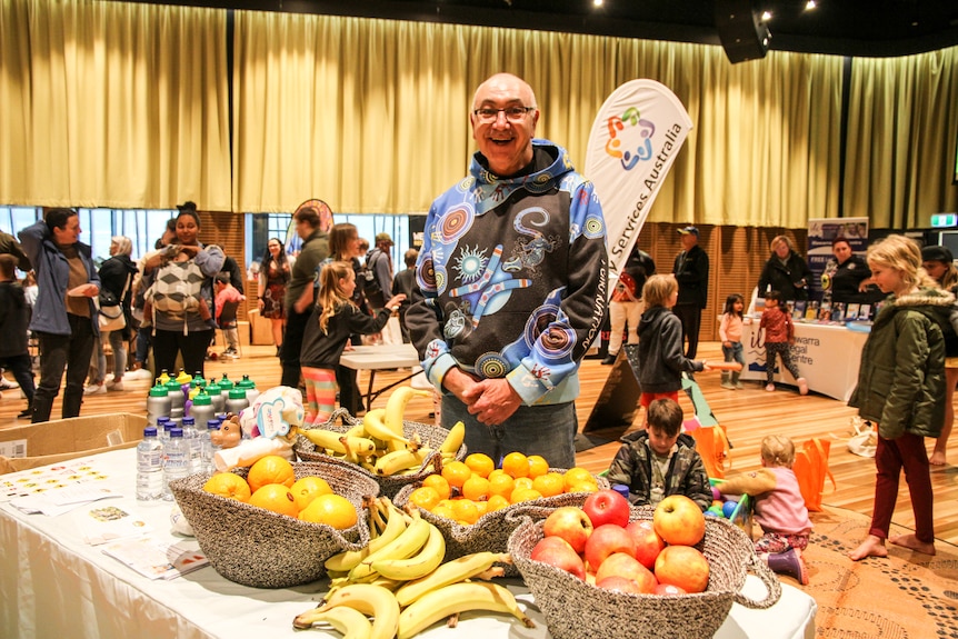 A man wearing a brightly-coloured jumper stands behind a table covered with baskets of fruit