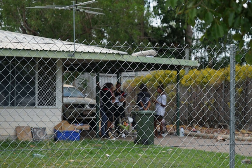 Young kids are seen standing outside a home behind a fence.