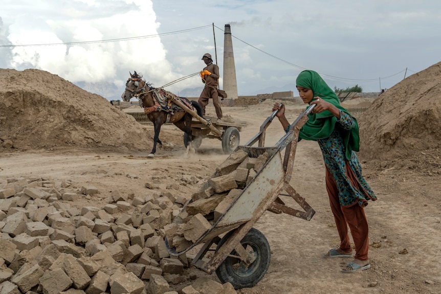 a young girl empties a wheelbarrow of bricks as a person rides a horse and cart behind her