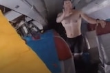 A man wearing a pair of black shorts and no shirt stands in the aisle of a half sunk plane, water laps at the seats' armrests