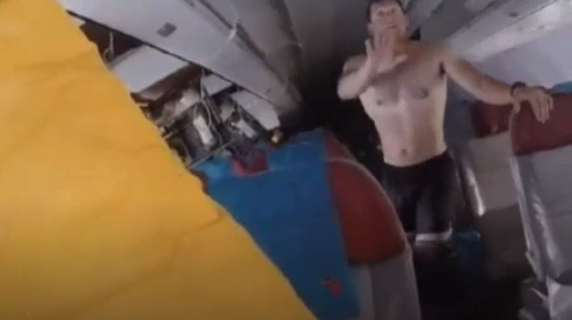 A man wearing a pair of black shorts and no shirt stands in the aisle of a half sunk plane, water laps at the seats' armrests
