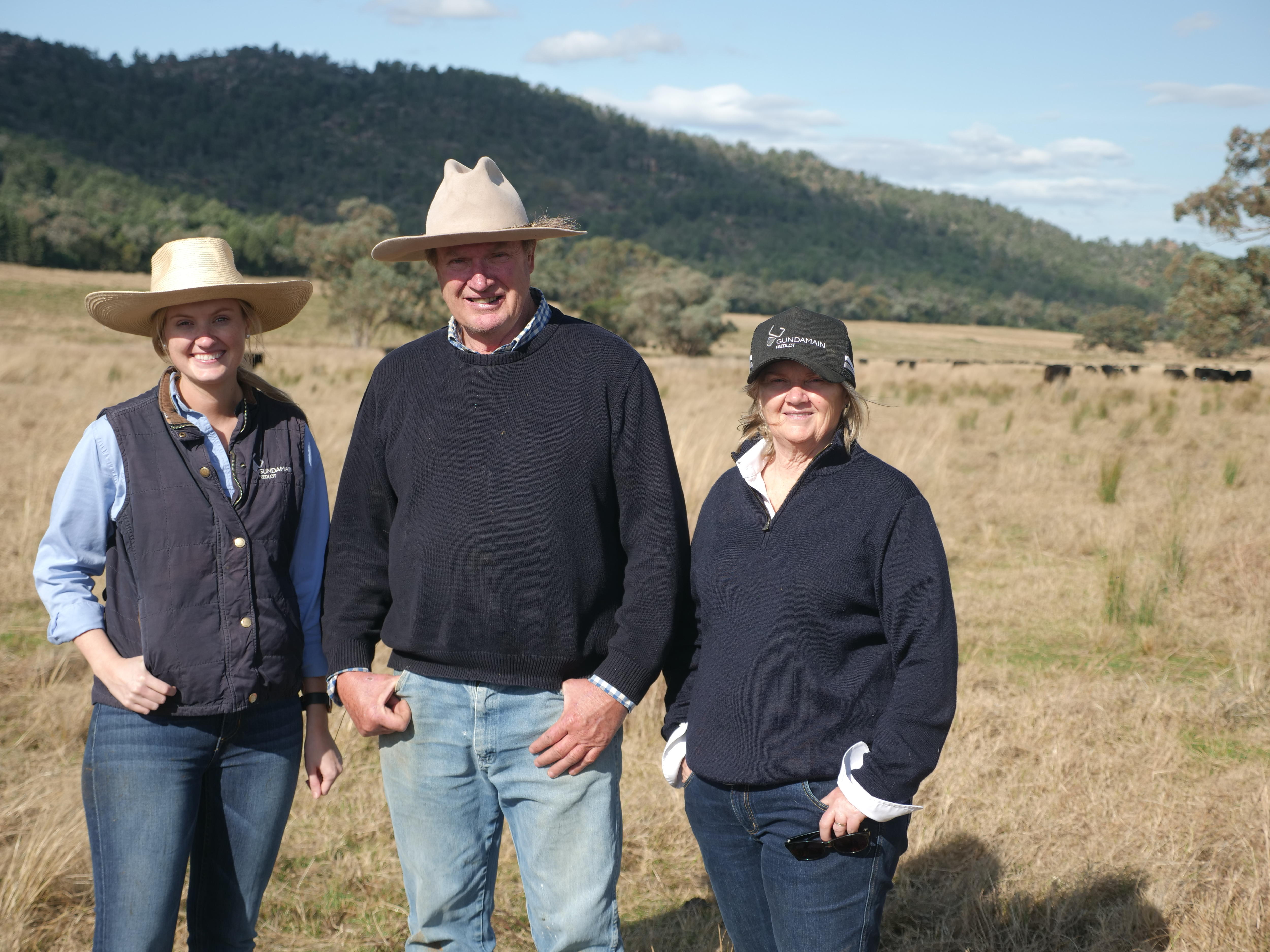 A young woman with an older man and woman all wearing hat in paddock with cattle behind.
