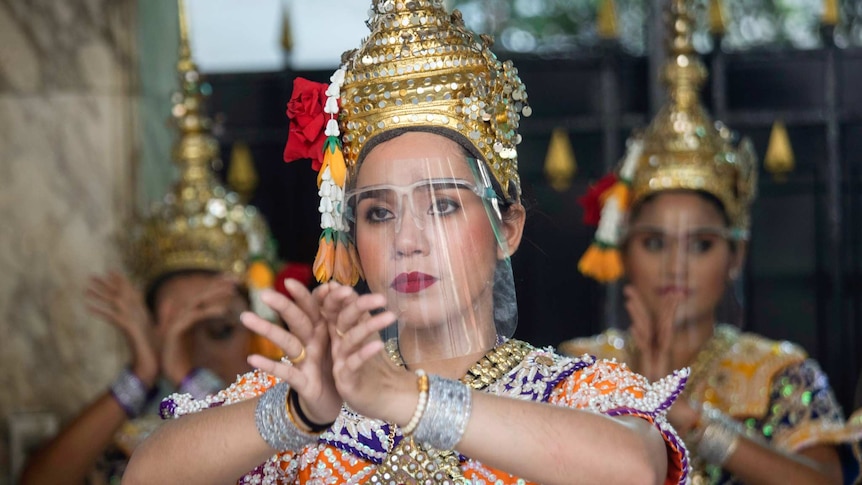 Three women wearing face shields, gold headwear and colourful costumes hold their hands up