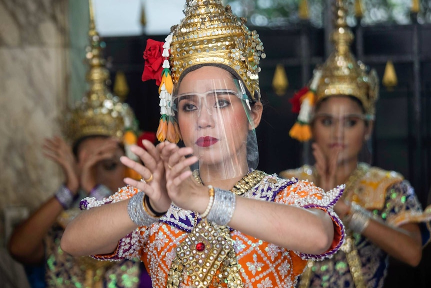 Three women wearing face shields, gold headwear and colourful costumes hold their hands up