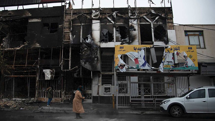 A woman wearing a coat and scarf stands in front of a burnt-out building