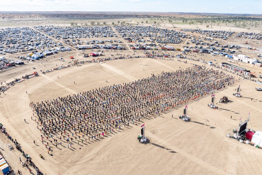 An aerial shot of people lined up on a dusty field, with a car park surrounding them. 