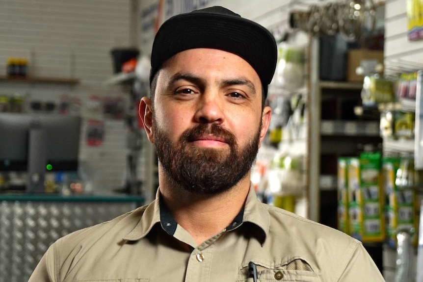 Alex Taskun in khaki shirt and black baseball cap standing in a workshop