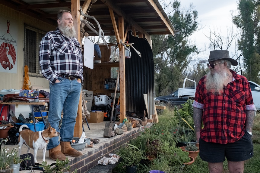 Two men with long beards stand by a shed with a deck