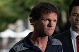 A man with clenched jaw holds his hands behind his back while addressing media.