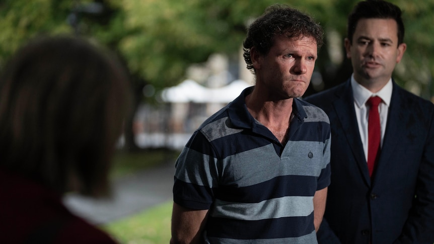 A man with clenched jaw holds his hands behind his back while addressing media.