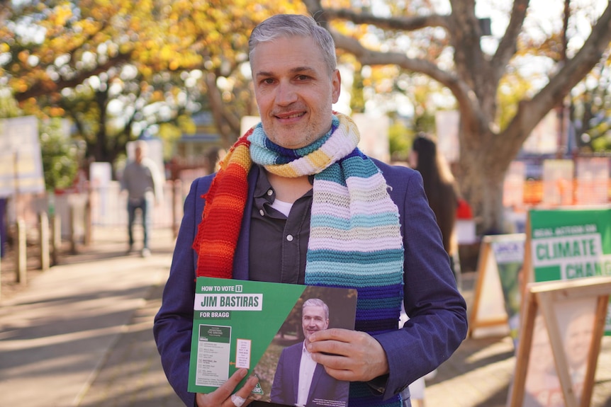 A man with a beard wearing a colourful scarf while holding a how to vote card