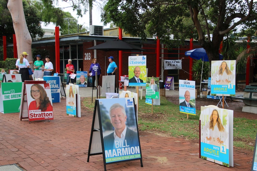 A group of volunteers wearing election campaign t-shirts stands outside a voting centre surrounded by election campaign posters.