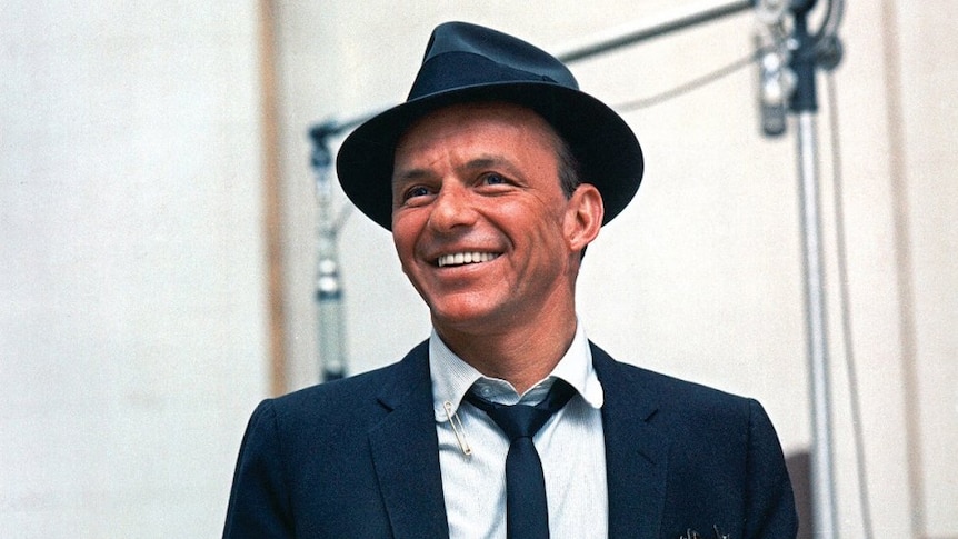 Singer Frank Sinatra stands smiling in a recording studio, wearing a black hat and suit