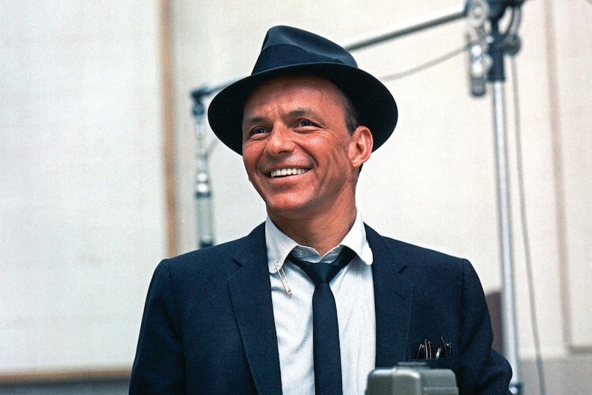 Singer Frank Sinatra stands smiling in a recording studio, wearing a black hat and suit