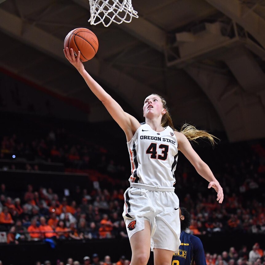 A girl wearing a white uniform playing basketball, shoots an orange basketball at the hoop in front of a crowd