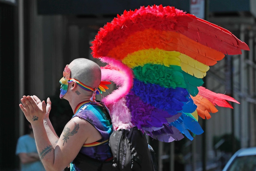 A man wearing rainbow wings and a rainbow coloured mask claps as he participates in a queer liberation march in New York.