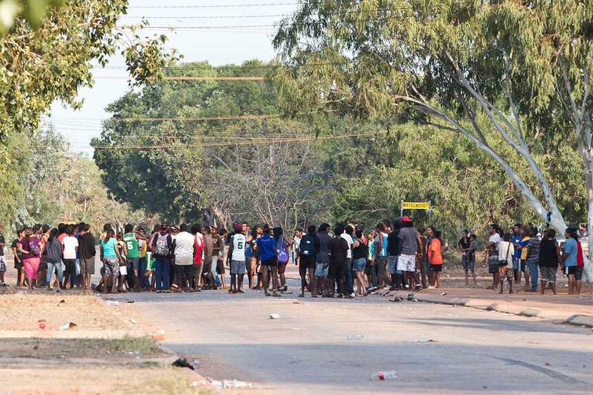 A large crowd of Indigenous people form on a street covered with rubbish