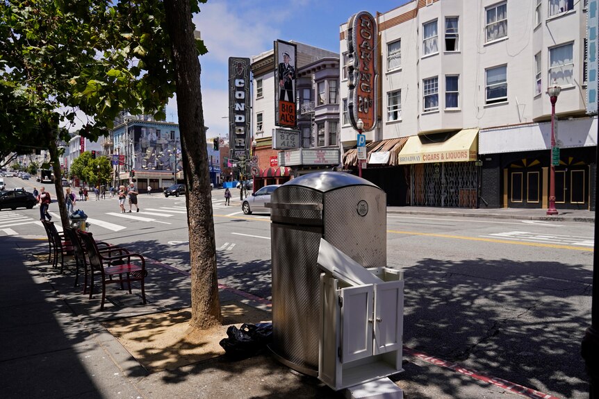 A curved, squat metal garbage bin sits on the side of a San Francisco street. A broken cabinet rests against it.