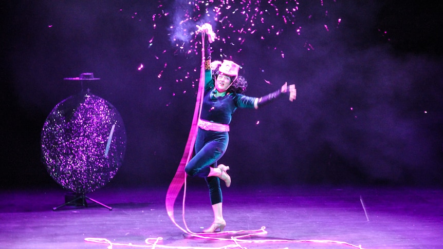 A woman in a purple velvet pantsuit dances with a pink ribbon on stage in front of a glitter ball.