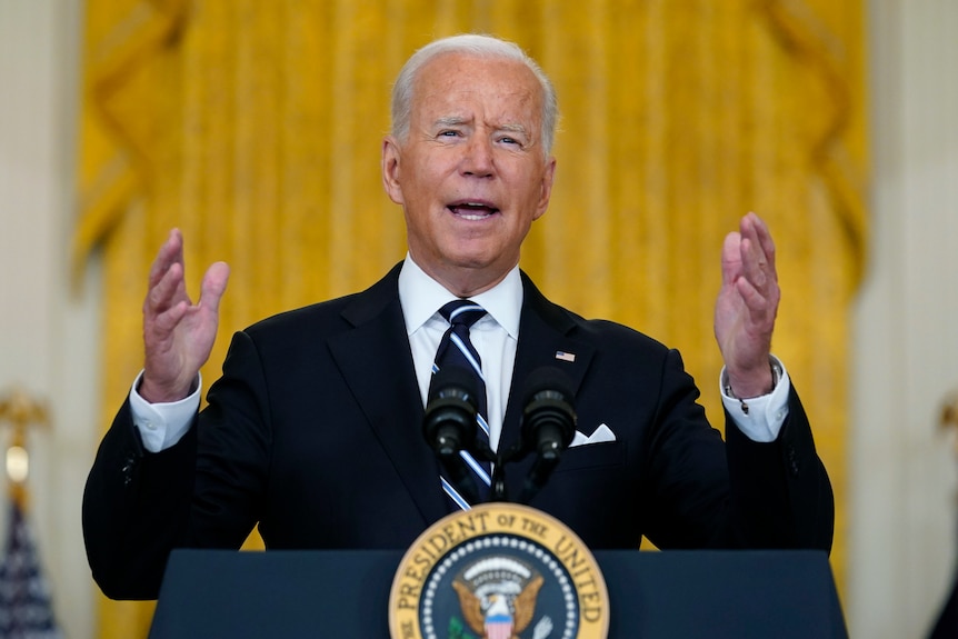 Joe Biden dressed in a suit stands at a podium infront of a yellow background.