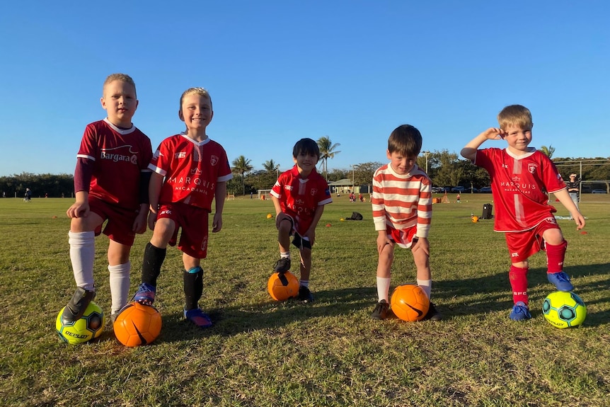 5 young boys wear soccer uniforms and have balls under their feet.