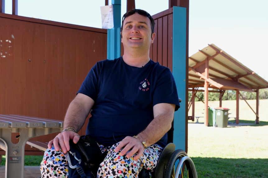 a man sitting in a wheelchair smiles at the camera, he's sitting underneath a barbecue shelter