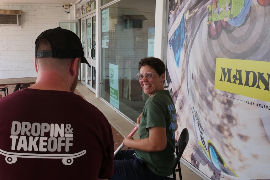 A man in a cap and maroon tshirt talks to a woman with short hair, glasses and a green shirt.