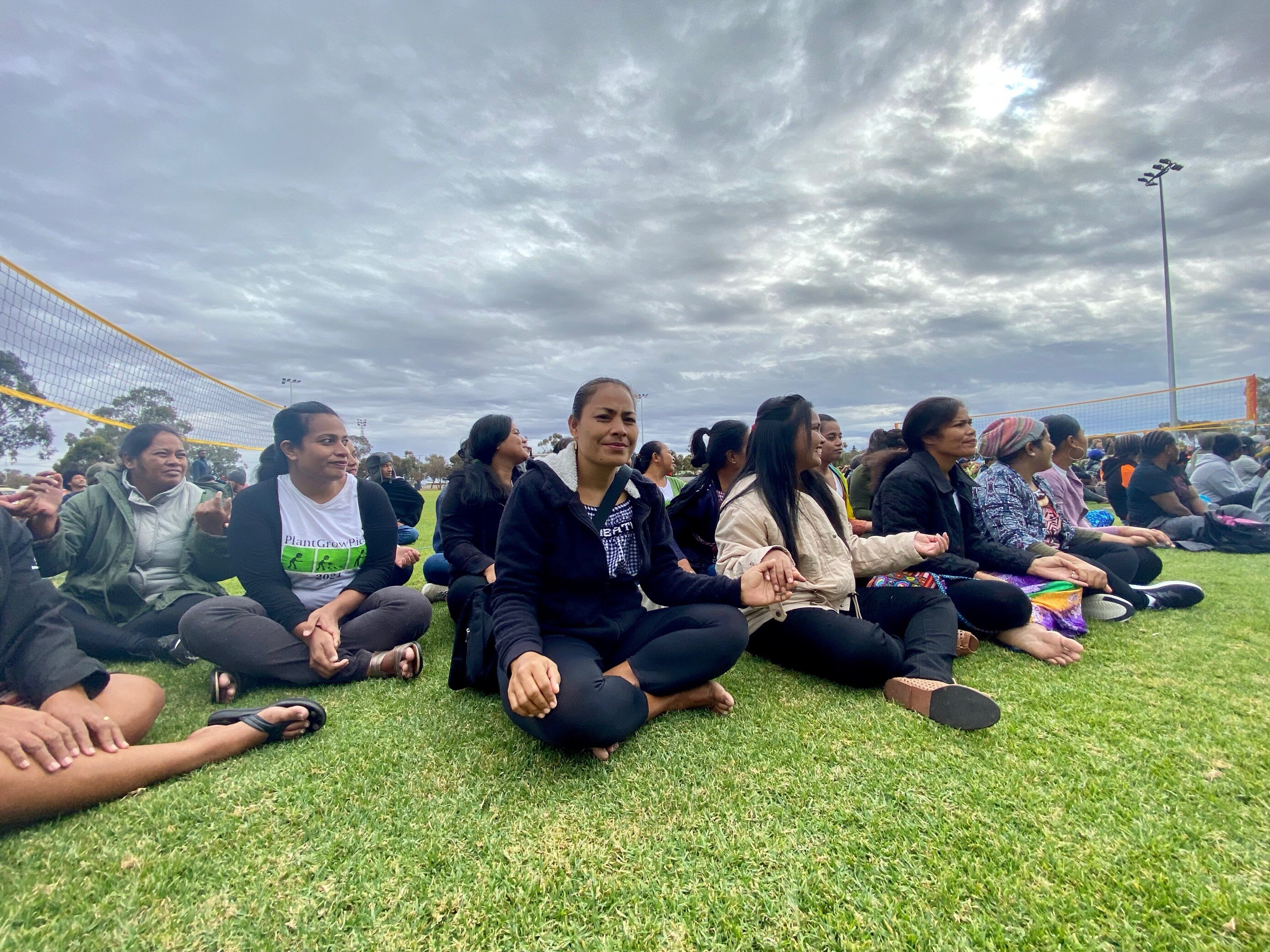 A large group of Pacific Islander women sit, crossed-legged on the grass watching the competition