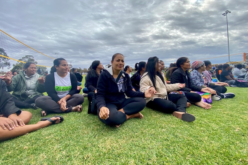 A large group of Pacific Islander women sit, crossed-legged on the grass watching the competition