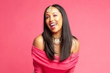 A woman wearing a pink top and white skirt stands in front of a bright pink wall, smiling