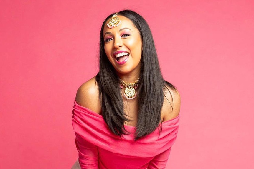 A woman wearing a pink top and white skirt stands in front of a bright pink wall, smiling