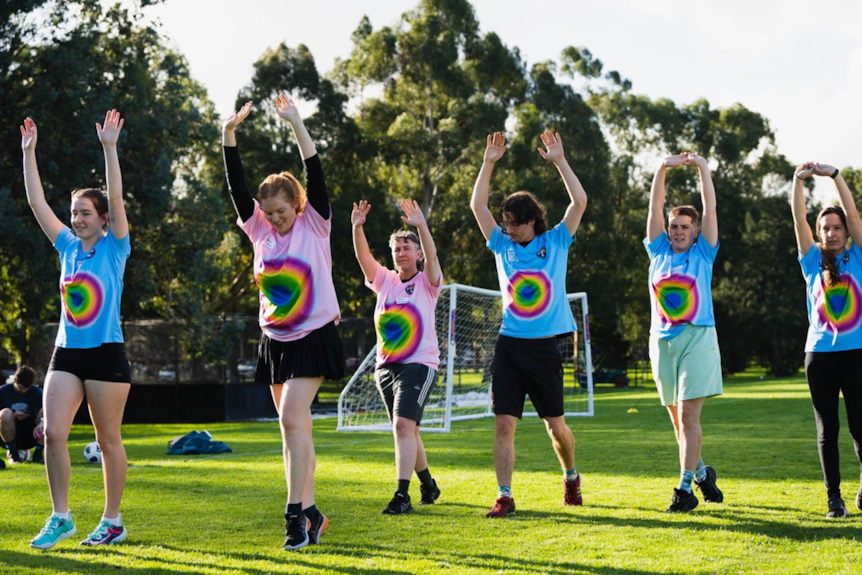 Six people wearing t-shirts and shorts have their arms raised above their heads, as they warm up for a football match.