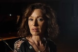 Tina Arena stands looking at the camera on a stage with dramatic lighting and a piano in the background.