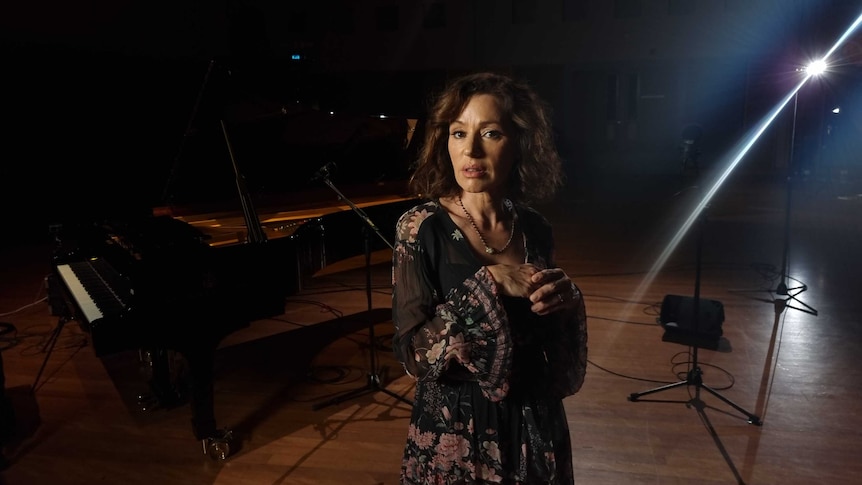 Tina Arena stands looking at the camera on a stage with dramatic lighting and a piano in the background.