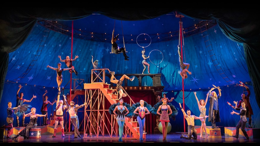 Lit stage showing circus scene, with cast of performers in various poses.