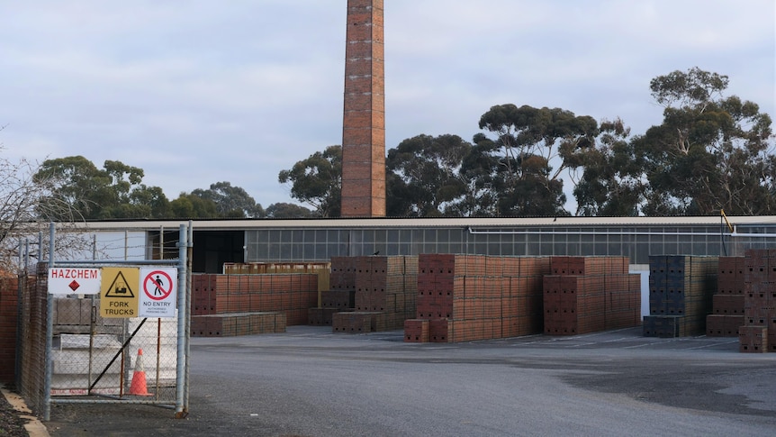 A industrial site showing stacked pallets of red bricks with a brick chimney behind. Steel gates with do not enter signs on side