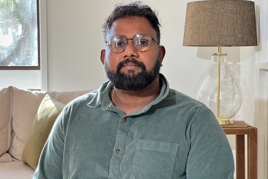 Supun Pollgolla looks seriously into the camera, sitting in a living room. He has a beard and wears glasses.