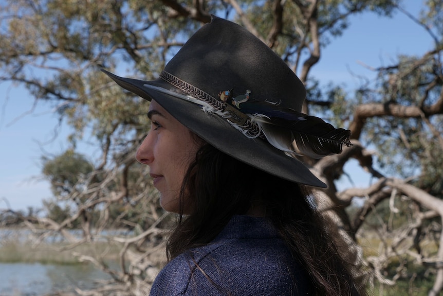 A woman with dark brown hair wears a felt hat with bird feathers and badges. She looks over a bed of water.