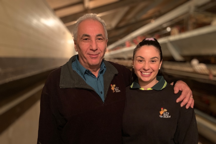 An older man stands in a poultry shed with his arm around a smiling dark-haired woman.