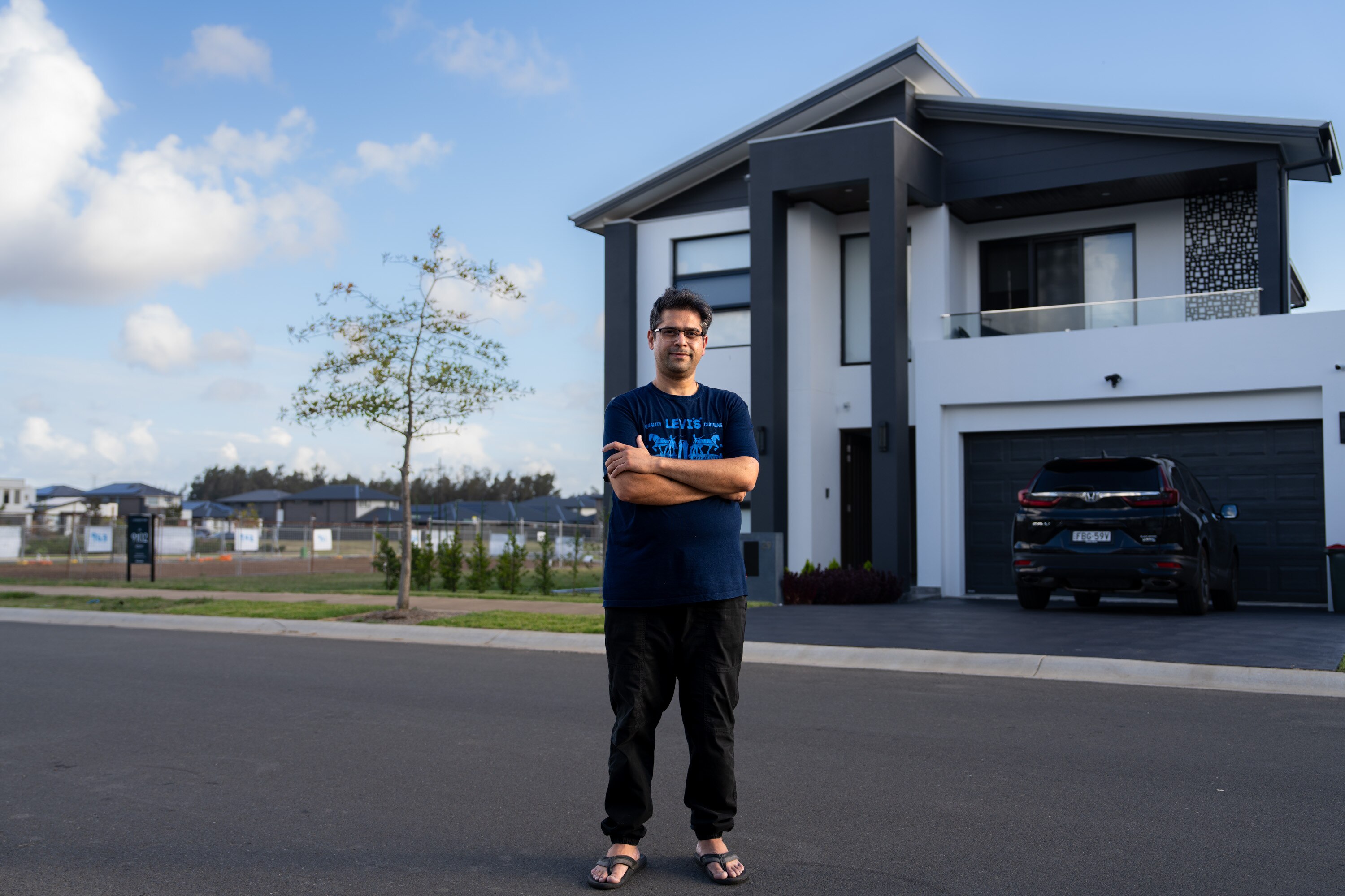 A man stands with his arms crossed in front of a house next to an empty lot. 