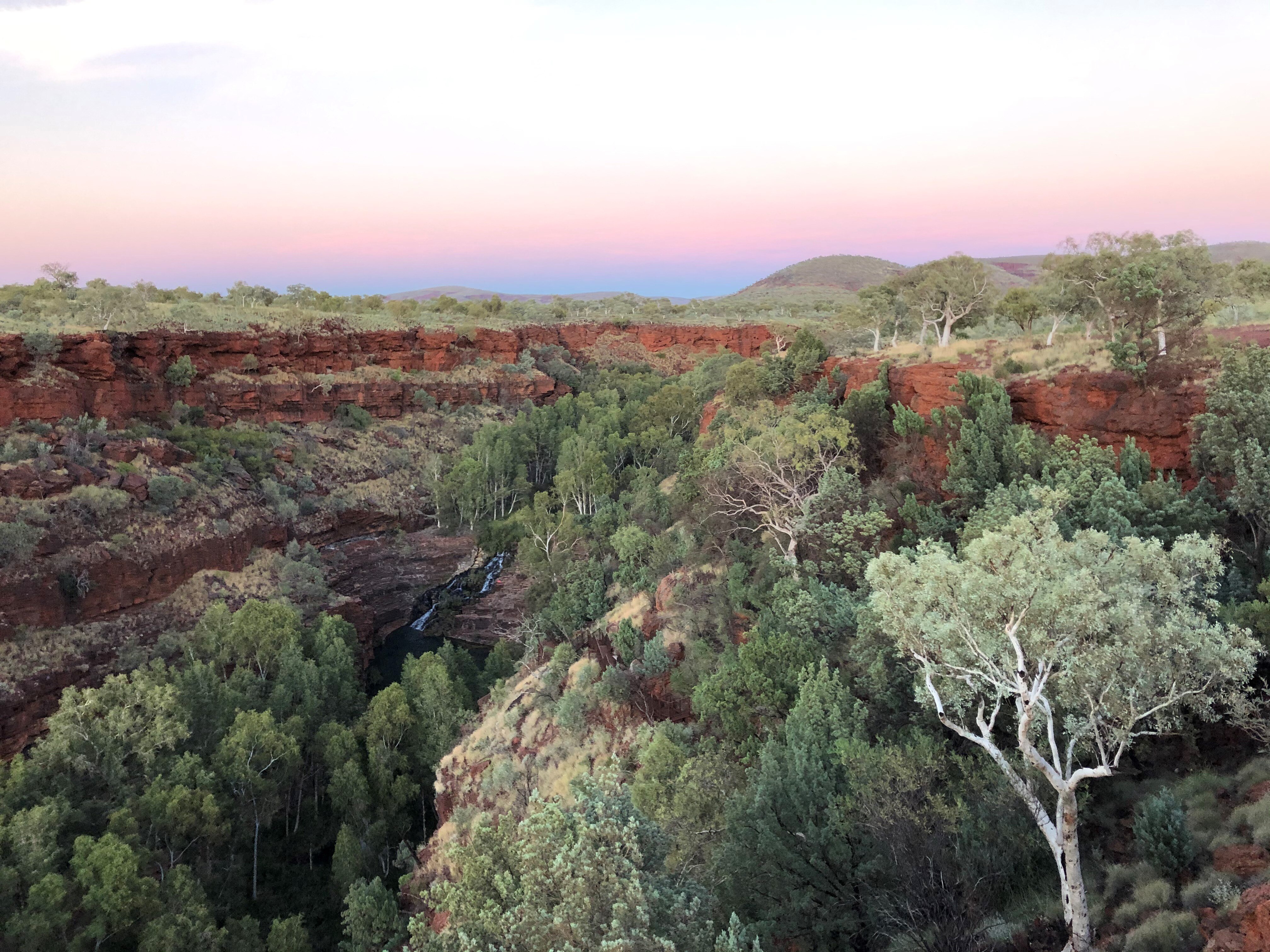 Large view of a red rock gorge with waterfall through the centre and sunset in the distance. 