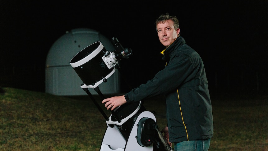 A man holding a telescope on a hill against a dark night sky. 