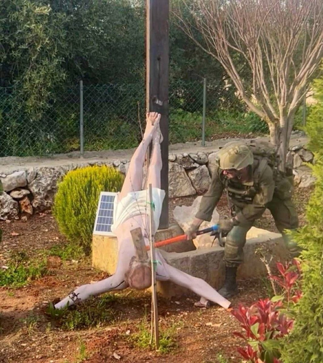 A soldier in uniform swings the blunt side of an axe at a fallen sculpture of Jesus on a corss