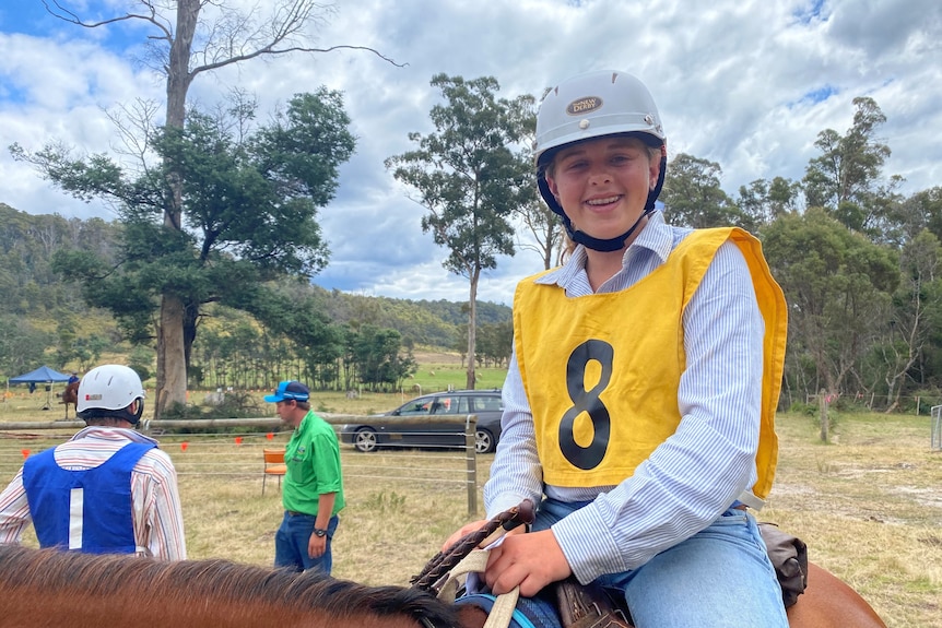 A young woman in a yellow bib sits on a chestnut horse, a man with a blue bib and a man in a green shirt stand behind