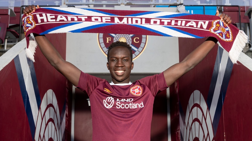 Garang Kuol holds up a Heart of Midlothian scarf while standing in the tunnel at the home ground of the football club.
