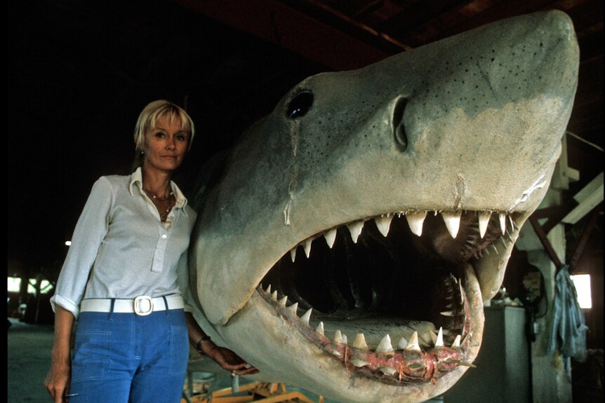 Archival photo of blonde-haired woman in jeans and white shirt standing beside giant model of shark's head.