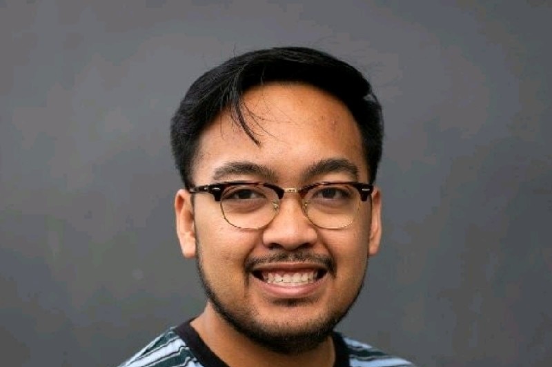 A young man with dark hair and glasses smiles at a camera while wearing a black and blue striped tshirt