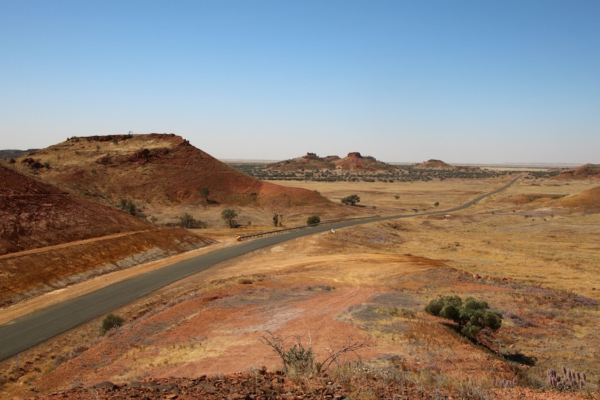 A bitumen road winds through vast, stark red yellow outback landscape and hills.