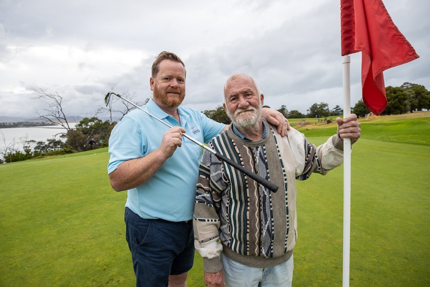 Two men stand on a golfing green, one holding a golf club, the other holding a golf flagstick
