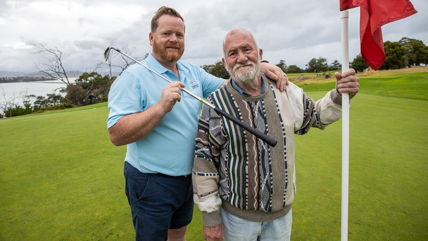 Two men stand on a golfing green, one holding a golf club, the other holding a golf flagstick